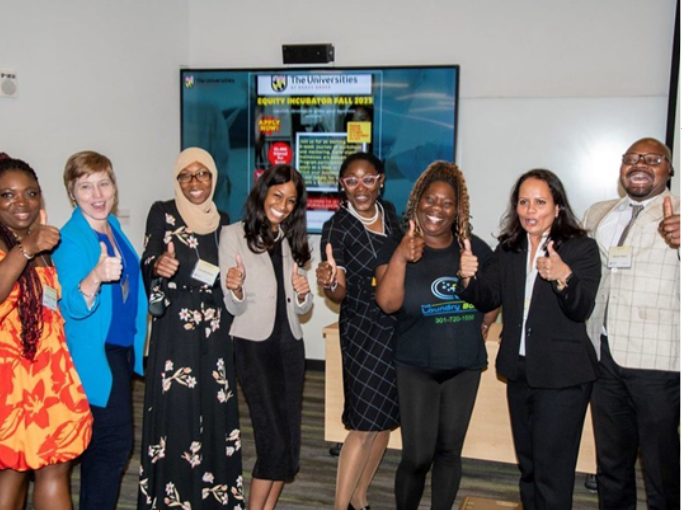 A diverse group of eight people smiling and giving thumbs up. They're in a room with a screen showing a presentation about the Equity Incubator Fall 2023.