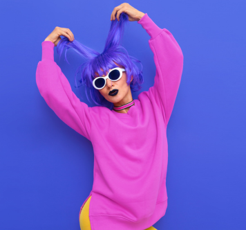 A woman in a vibrant pink sweater strikes a playful pose against a blue background, wearing a blue wig, white sunglasses, and bold lipstick.