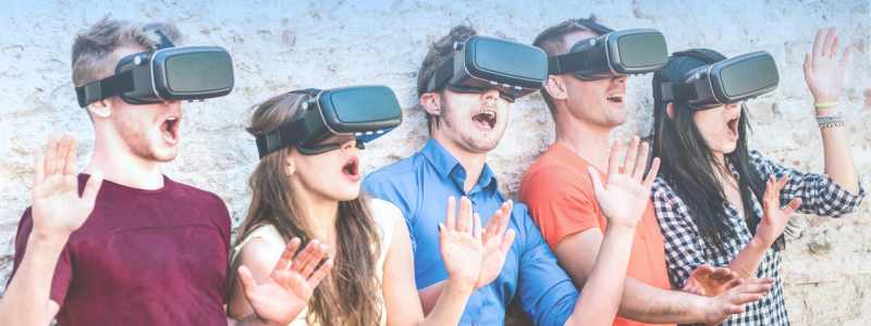 A group of five people wearing VR headsets, standing against a light brick wall, appear amazed and excited, with their hands raised in surprise.