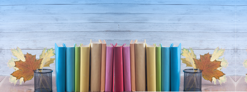 Books with colorful covers stand upright on a table against a blue wooden background. Fall leaves are placed in desk organizers on either side.