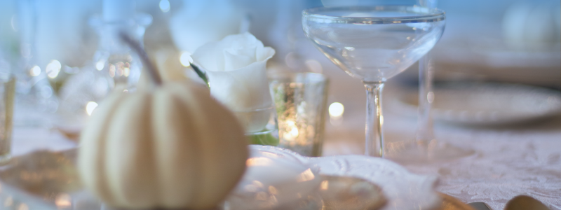 Elegant table setting with a small white pumpkin, a white rose in a glass, and a translucent coupe glass, all softly lit for a serene ambiance.