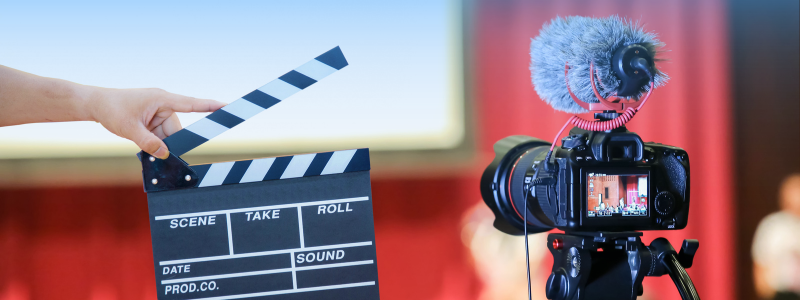 A hand holds a film clapperboard beside a camera with a microphone on a tripod. The background has red curtains.