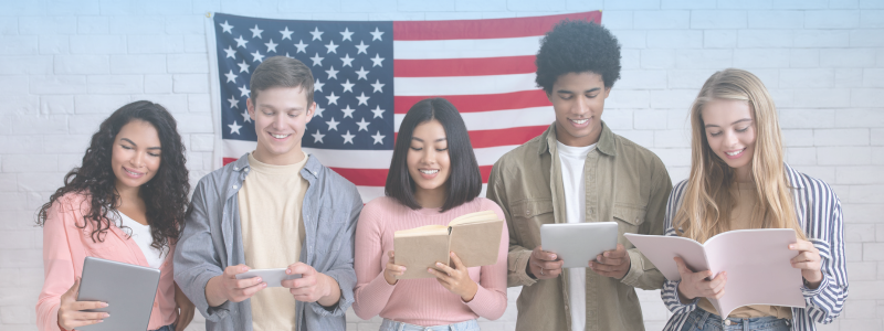 Five diverse young adults reading books and digital devices, stand smiling in front of an American flag.
