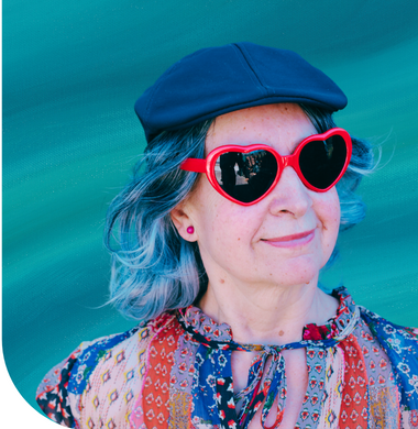 A woman with blue hair smiles, wearing red heart-shaped sunglasses and a black cap. Her colorful patterned shirt contrasts with the teal background.