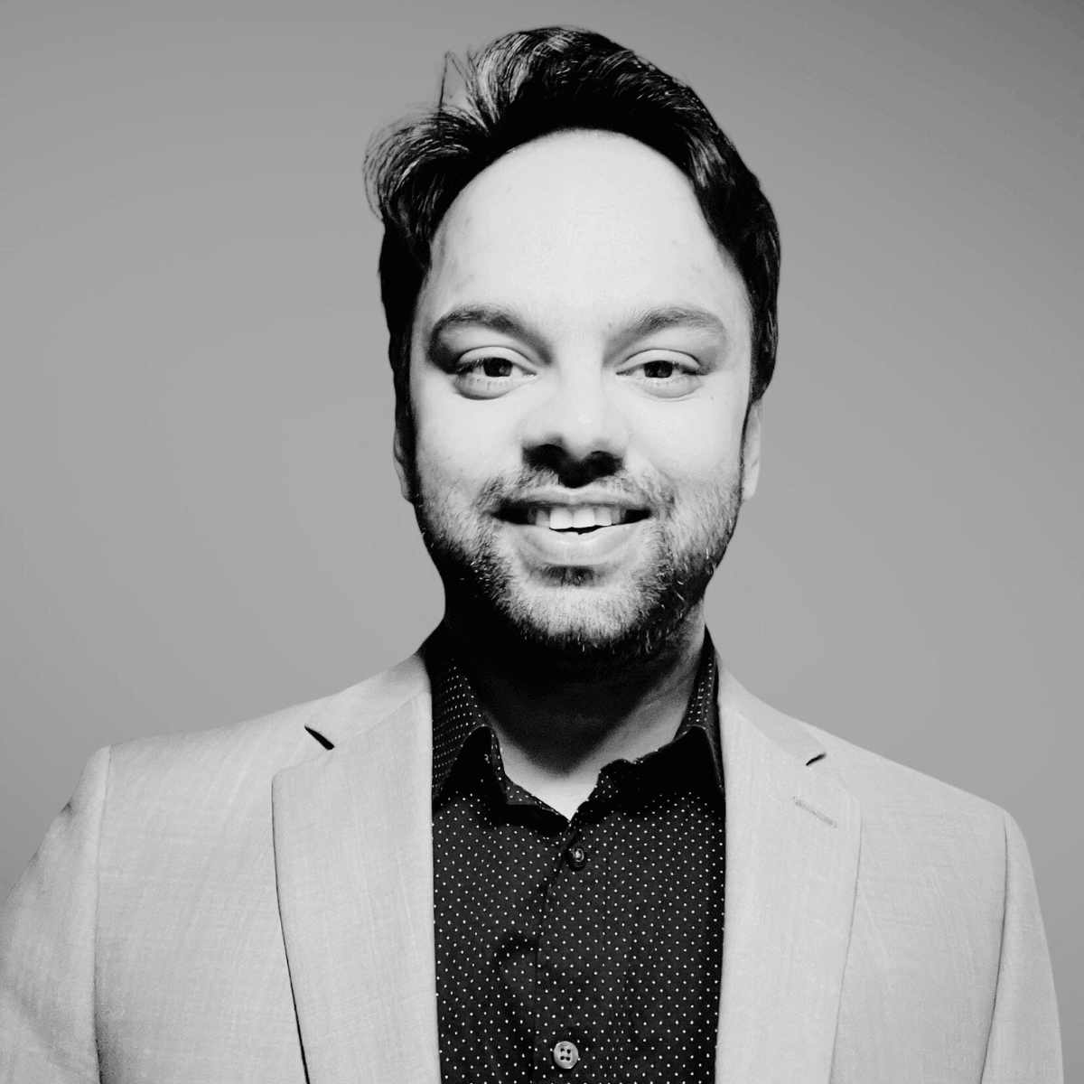 Smiling man in a light blazer and polka dot shirt against a plain background. His confident expression conveys a friendly and approachable tone.