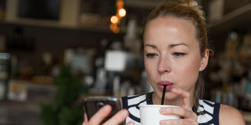 oct-2023-J.pngA woman in a striped top sips from a cup with a straw while focusing on her phone in a cozy cafe. The background is blurred, creating a relaxed atmosphere.