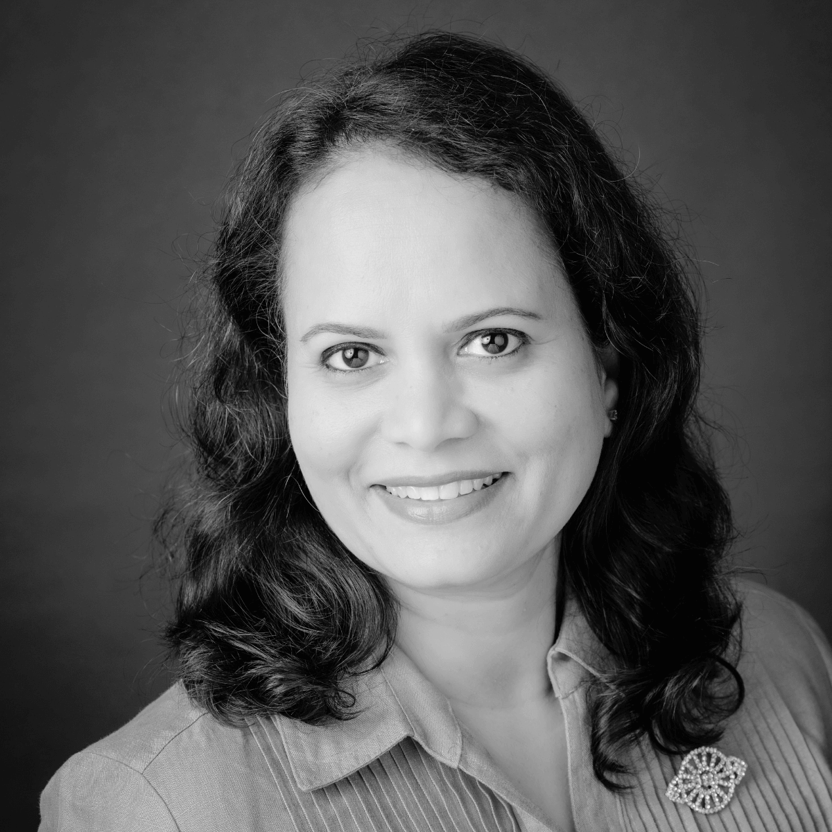 Black and white portrait of a woman with wavy hair smiling warmly. She wears a blazer with a decorative brooch. Neutral background adds elegance.